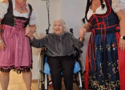 2 lächelnde Damen nehmen Seniorin im Rollstuhl liebevoll an die Hände